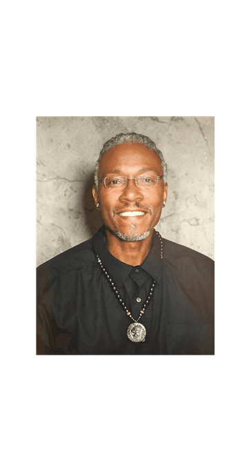 Smiling Black man with gray hair, glasses, and lion pendant necklace against gray wall.